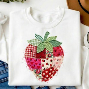 May include: White sweatshirt featuring a large, patchwork strawberry design. The strawberry is composed of various fabric patterns, including red gingham, floral, and heart prints. Green leaves and stem complete the applique. The sweatshirt is a casual, comfortable garment.