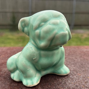 May include: A light green ceramic bulldog figurine. The bulldog is sitting with its front paws extended and its head tilted slightly to the side. The figurine has a glossy finish and is a solid color.