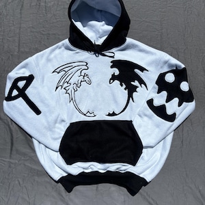 May include: White hoodie with black detailing, showcasing dragon and axe motifs. The hoodie has a black hood, pocket, and cuffs. Dragon silhouettes are embroidered on the front, with an axe on the left sleeve and a mushroom design on the right sleeve.