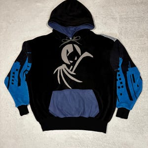 May include: A black hooded sweatshirt featuring a light grey graphic of a winged figure centred on the chest. The sleeves display a blue cityscape pattern, while the front pocket and hood lining are a muted blue. It has a drawstring hood.