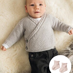 May include: A baby wearing a light gray, wrap-style knit sweater with two dark buttons. The baby is also wearing black corduroy pants and a pair of light pink knit baby socks. The baby is lying on a white, fluffy surface.