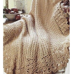 May include: A beige crochet blanket featuring a textured, repeating stitch pattern throughout the main body and a wide, decorative scalloped border along the edges. The fabric is crafted from yarn in a neutral tan shade with a visible openwork design.