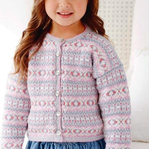 May include: A young child wearing a button-up cardigan. The sweater is knit with a pattern of pink, gray, and white geometric shapes. The child is also wearing a blue denim skirt. The cardigan has a round neckline and long sleeves.