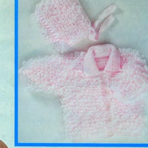 Pode incluir: Um conjunto de bebê de malha rosa claro. O conjunto inclui um casaco com gola, um gorro e um par de luvas. A textura de malha é visível em todo o conjunto, criando uma aparência macia e aconchegante.