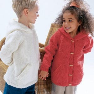Op de afbeelding: Twee kinderen dragen gebreide truien met capuchon. Eén is wit, de andere rood. Beide hebben een voorzak en houten knopen. De kinderen houden elkaars hand vast en glimlachen. De achtergrond omvat een rieten stoel.