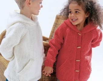 Op de afbeelding: Twee kinderen dragen gebreide truien met capuchon. Eén is wit, de andere rood. Beide hebben een voorzak en houten knopen. De kinderen houden elkaars hand vast en glimlachen. De achtergrond omvat een rieten stoel.