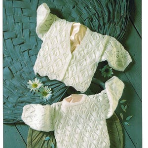 May include: Two white knitted baby sweaters with diamond and leaf patterns. One is a cardigan, and the other is a crew neck. The sweaters are displayed on a green surface with decorative leaves and flowers.