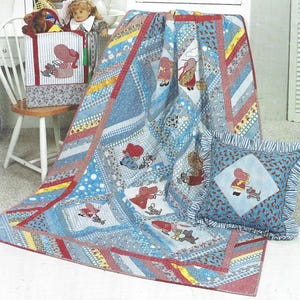 Puede incluir: Un juego de colcha, almohada y caja de almacenamiento de patchwork. La colcha presenta un patrón azul y blanco con detalles en rojo y amarillo, y figuras bordadas. La almohada tiene un borde con volantes. La caja de almacenamiento tiene un diseño a juego.