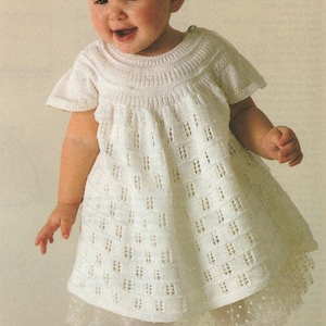 May include: A smiling baby wearing a white knitted dress with a textured pattern. The dress has short sleeves and a flared skirt, layered over a white dotted tulle skirt. The baby is also wearing white leggings and shoes.