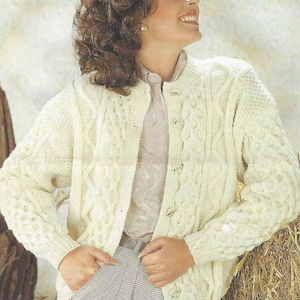 Puede incluir: Cárdigan de punto trenzado color crema con cierre de botones. El suéter presenta intrincados patrones tejidos y se combina con pantalones a cuadros grises y blancos. El estilo general es clásico y acogedor.