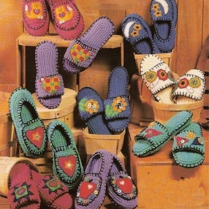 May include: A collection of colorful, handmade crochet slippers in various designs. The slippers feature heart, flower, and bear motifs in shades of pink, purple, blue, and green. They are displayed on wooden surfaces, showcasing the craft's cozy aesthetic.