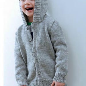 May include: A light gray knitted hooded cardigan with button closures. The sweater has a ribbed hem and cuffs. The child is wearing light blue jeans. The cardigan is a comfortable and stylish outerwear option for cooler weather.