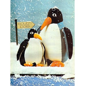Puede incluir: Dos figuras de pingüinos con cabezas negras, cuerpos blancos y picos y patas naranjas se encuentran en una escena nevada. Un letrero indica el "Polo Sur". El fondo es de color azul claro con motas blancas.