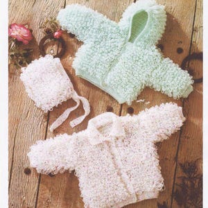 May include: Three pieces of baby clothing: a light green hooded jacket, a white jacket, and a white bonnet. The garments are made of a textured, looped yarn, and are displayed on a wooden surface. The bonnet has tie strings.