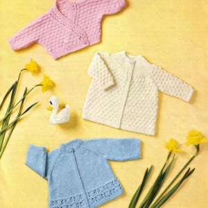May include: Three knitted baby sweaters in pastel colors: pink, cream, and blue. The pink sweater has a V-neck and button closure. The cream sweater is a classic design. The blue sweater features a decorative openwork hem. Yellow daffodils and a white duck figurine are also present.