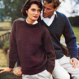 Puede incluir: Una pareja con jerséis de punto. La mujer lleva un jersey burdeos oscuro de cuello redondo sobre una camisa blanca con cuello. El hombre lleva un jersey azul marino de cuello en V sobre una camisa blanca con cuello.