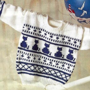 May include: White baby sweater with navy blue cat silhouettes and geometric patterns. The sweater has long sleeves with patterned cuffs and a round neckline. The design includes small buttons on the shoulder.