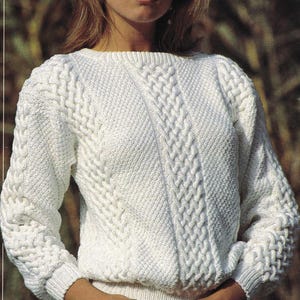 Könnte beinhalten: Ein weißer Zopfstrickpullover mit Rundhalsausschnitt und langen Ärmeln. Der Pullover hat ein strukturiertes Muster mit diagonalen und vertikalen Zopfmustern. Die Ärmel haben einen gerippten Bund. Der Pullover ist aus weichem, warmem Material.