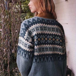 Op de afbeelding: Blauwe trui met een geometrisch patroon in wit, lichtblauw en geel. De trui heeft een ronde hals en lange mouwen. Het ontwerp bevat ruit- en bloemmotieven. De persoon draagt er een bruin overhemd met kraag onder.