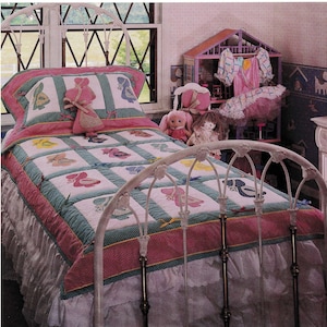 May include: A white metal bed frame with a floral design, dressed with a quilt featuring colourful shoe designs. The quilt has a pink and green border, with a ruffled white bed skirt. A doll and a dollhouse are visible in the background.