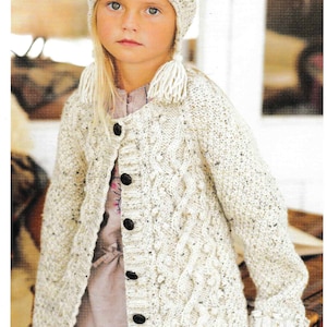 May include: A cream-colored knitted hat with a pom-pom and matching long cardigan with black buttons. The cardigan has a cable knit design and is worn by a young person.