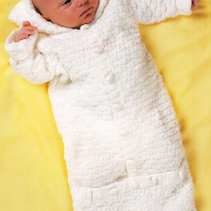 May include: A white, hooded baby sleeping bag with a textured knit pattern. The sleeping bag has a button closure down the front and small pockets. The baby is lying on a yellow surface.
