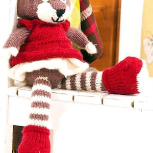 May include: A handmade knitted fox doll with a red dress and white trim. The fox has brown and white striped legs and red feet. The doll is sitting on a white surface. The fox has a brown head with white accents and black eyes.