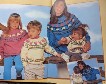 Op de afbeelding: Een familie draagt bijpassende gebreide truien. De truien zijn in tinten roze, crème en blauw, met decoratieve patronen van schapen en bloemmotieven. De kinderen dragen een blauwe broek en de volwassenen blauwe truien.