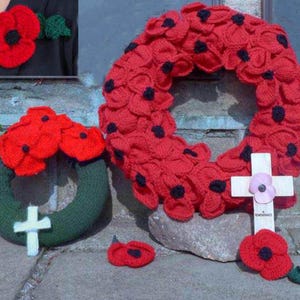 May include: Handmade red poppy wreaths and a poppy pin with a green leaf. The wreaths are made of red knitted poppies with black centers. One wreath is on a green base with a white cross. A white cross with a pink poppy is also present.