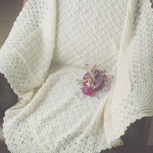 May include: Ivory-colored knitted blanket with a diamond pattern and a scalloped lace edge. A small bouquet of pink and purple flowers rests on the blanket. The blanket is draped over a chair.