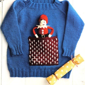 Op de afbeelding: Een blauwe gebreide trui met een rood en wit zakontwerp. Een kleine pop met een rode hoed en jas zit in de zak. Een gele cracker met witte sterren ligt rechts van de trui.