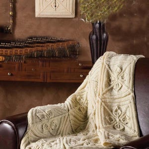 May include: Cream-colored knitted blanket draped over a brown leather armchair. The blanket features geometric and floral patterns. A matching knitted clock hangs on the wall. A dark vase with greenery and antique key are also visible.