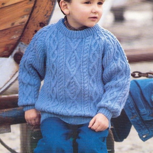 Può includere: Un bambino con un maglione a trecce blu chiaro e jeans blu. Il maglione presenta un motivo dettagliato e maniche lunghe. Il bambino è seduto vicino a una barca di legno.