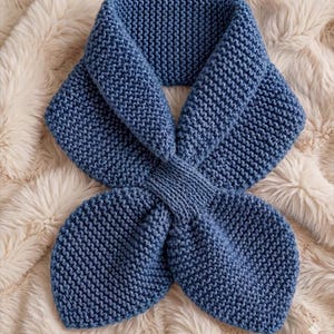 May include: A blue knitted scarf featuring a garter stitch texture and a central crossover design. The scarf is constructed with a wide collar section that tapers into a knotted middle, creating two rounded ends with a consistent blue yarn.