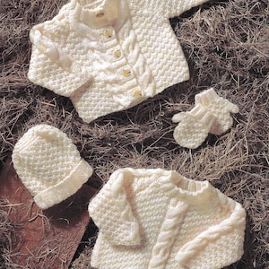 May include: Cream-colored knitted baby clothing set, including two sweaters, a hat, and mittens. The sweaters have button closures and cable knit detailing. The set is displayed on a textured, brown surface.