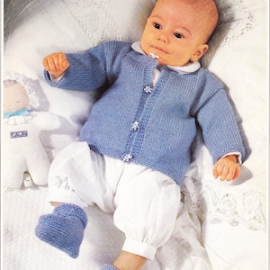 May include: A baby wearing a light blue knitted cardigan with white buttons and a white collar, paired with white bloomers and matching blue booties. A white doll with a blue bow is on the left.