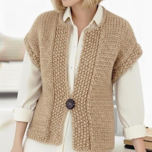 Puede incluir: Un chaleco de punto color beige con un cierre de botón grande de color marrón oscuro. El chaleco tiene un cuello redondo y mangas cortas. La modelo lleva el chaleco sobre una camisa blanca abotonada.