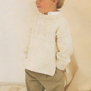 Puede incluir: Un niño viste un suéter de punto crema con un diseño texturizado en el pecho y pantalones caqui. El suéter tiene un cuello alto y mangas largas. El niño está de pie con las manos en los bolsillos.