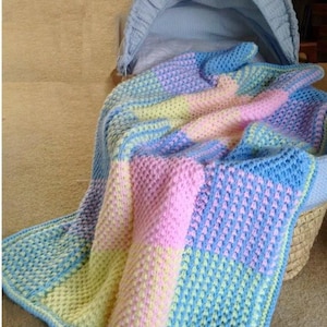 Op de afbeelding: Een handgemaakte, pastelkleurige babydeken in een wieg. De deken heeft een gestructureerd steekpatroon en is voornamelijk roze, geel en blauw. De wieg is lichtblauw en gemaakt van geweven materiaal.