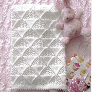 May include: A white knitted baby blanket with a scalloped edge and a geometric pattern. A pink stuffed bunny and a wrapped gift with a cupcake design are next to the blanket. The background is a pink knitted fabric.