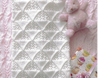 May include: A white knitted blanket featuring a geometric triangular pattern with textured sections. A small, light pink plush rabbit toy is positioned near the top right corner, and a gift wrapped in cupcake-patterned paper sits at the bottom right.