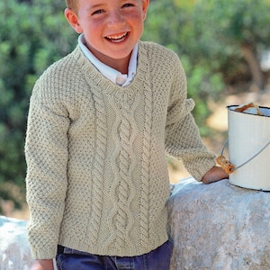 May include: A light beige knitted sweater with a cable knit design down the center and a textured pattern on the sleeves. The sweater has a V-neck and is worn over a white collared shirt. The person is wearing blue jeans.