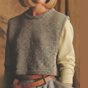 May include: A sleeveless, cropped, heathered grey knit sweater vest with a round neckline and ribbed trim, worn over a long-sleeved, solid pale yellow shirt, paired with a brown leather belt featuring a rectangular buckle and a grey textured skirt.
