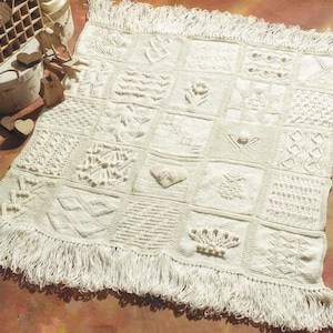 May include: Cream-colored, square-patterned knitted blanket with a fringed edge. The blanket features various textured squares with different designs, including floral and geometric patterns. The blanket is displayed on a brown surface.