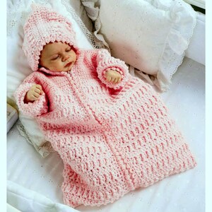 May include: A baby wearing a light pink crocheted outfit, including a hooded jacket and matching blanket. The outfit is made of soft yarn and has a textured pattern. The baby is lying on a white surface with a pillow in the background. The overall theme is baby clothing.