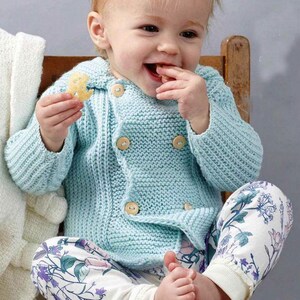 Può includere: Un bambino indossa un cardigan lavorato a maglia azzurro con bottoni in legno e pantaloni con stampa floreale. Il bambino è seduto su una sedia di legno e sorride mentre tiene uno spuntino.