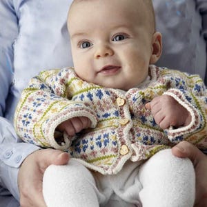 Puede incluir: Un bebé sonriente con una rebeca de punto estampada con botones de madera en forma de corazón. La rebeca presenta una mezcla de diseños florales azules, amarillos, rosas y verdes. El bebé también lleva pantalones blancos.