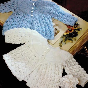 May include: Two knitted baby cardigans. One is light blue with a textured pattern and white buttons. The other is white with a similar design. Both have a round collar and long sleeves. The cardigans are displayed on a vintage book.