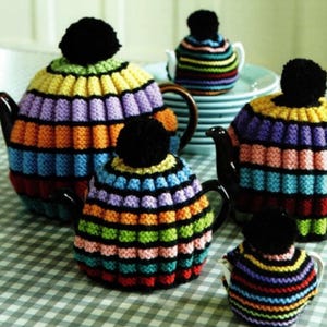 Könnte beinhalten: Eine Ansammlung von Teekannen mit bunten, gestreiften Strickhüllen. Die Hüllen weisen eine Vielzahl von leuchtenden Farben auf, darunter Gelb, Lila, Orange und Grün, mit einem schwarzen Bommel oben. Die Teekannen stehen auf einer karierten Oberfläche.
