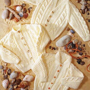 Puede incluir: Cuatro cárdigans de punto crema hechos a mano con cierres de botones. Los suéteres presentan patrones de punto trenzado y texturizados. Las prendas se exhiben sobre una superficie arenosa con conchas marinas y guijarros.